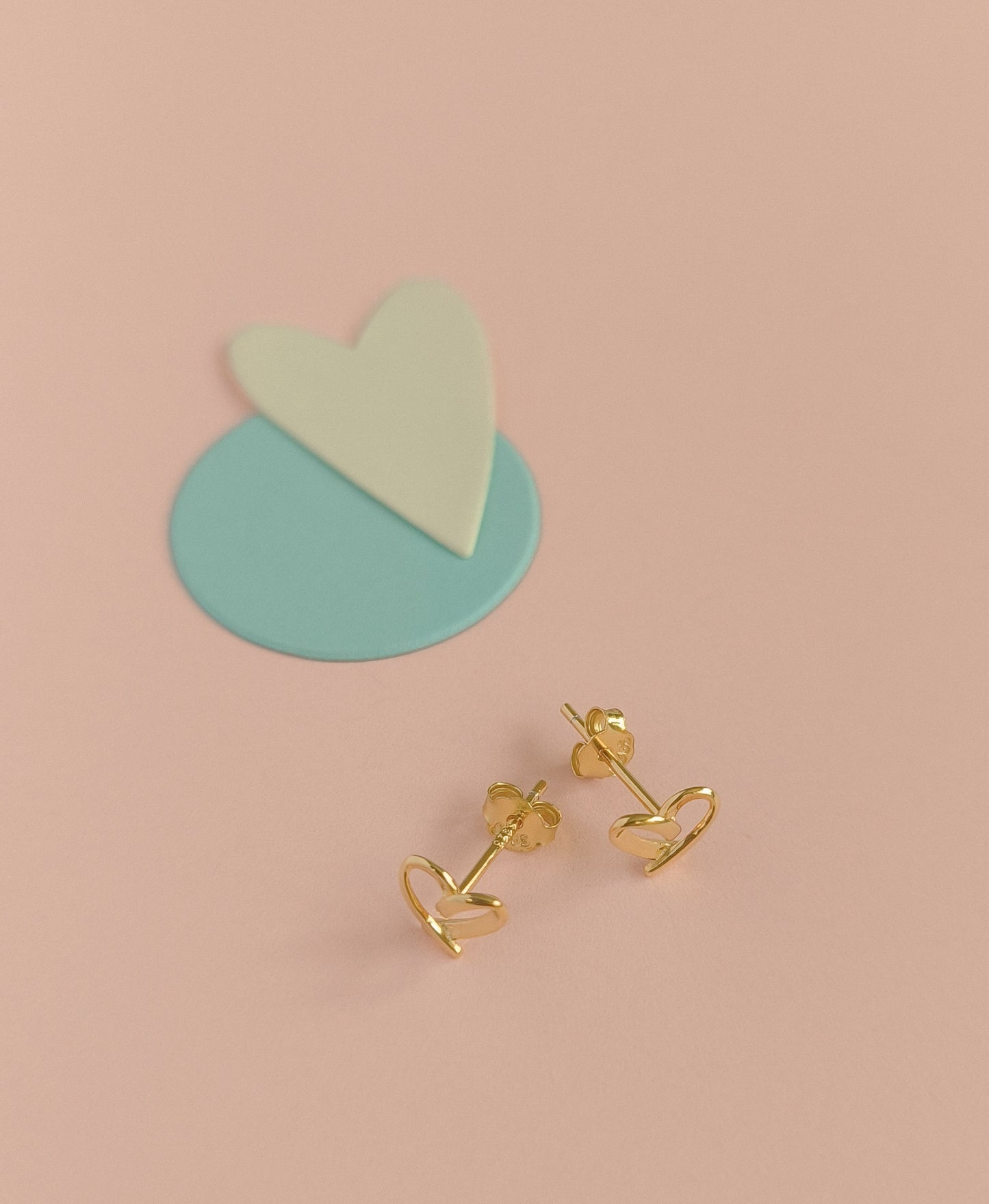 Gold heart-shaped earrings on a pink background with a heart-shaped stand.