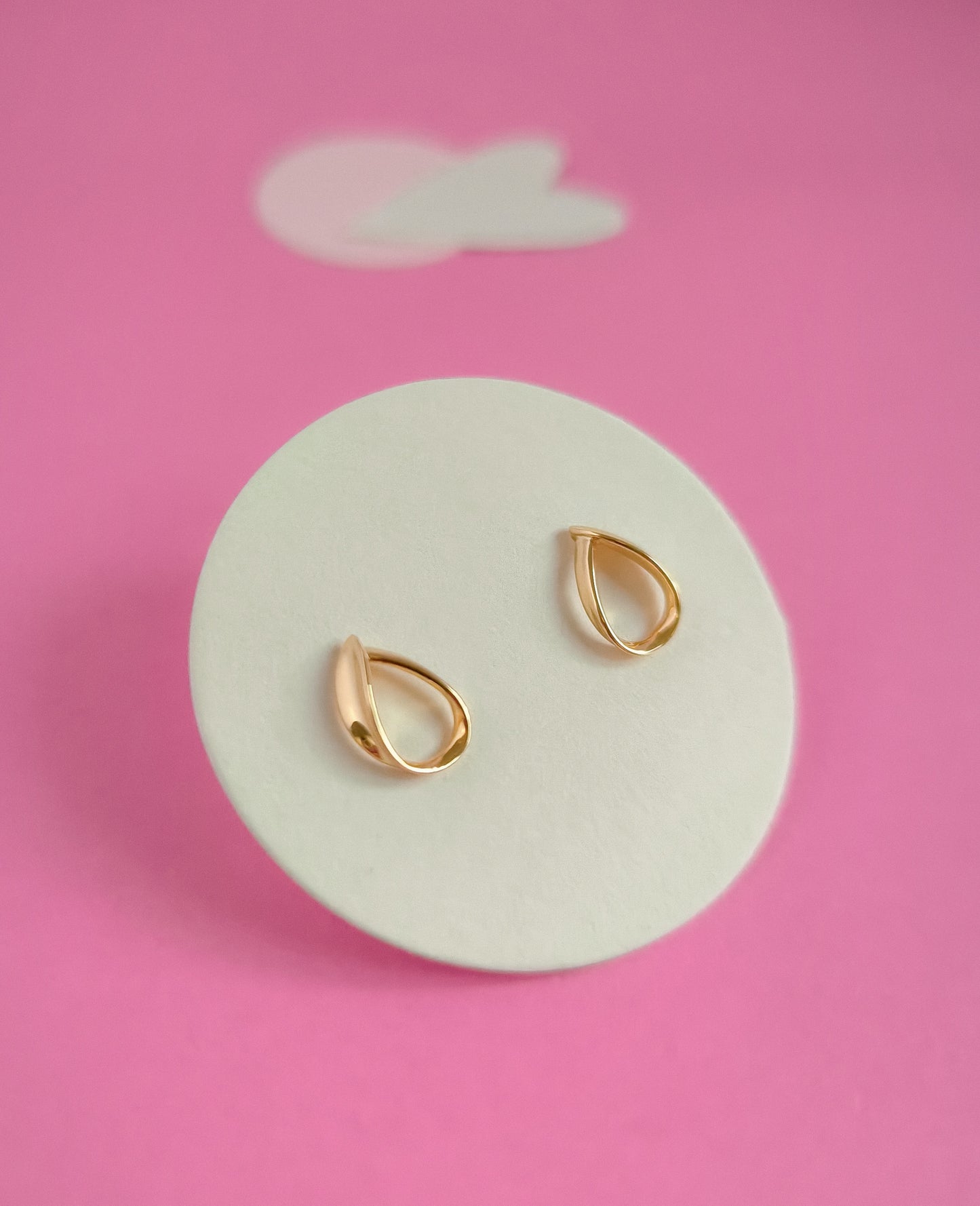 Gold teardrop earrings on a white circular stand against a pink background