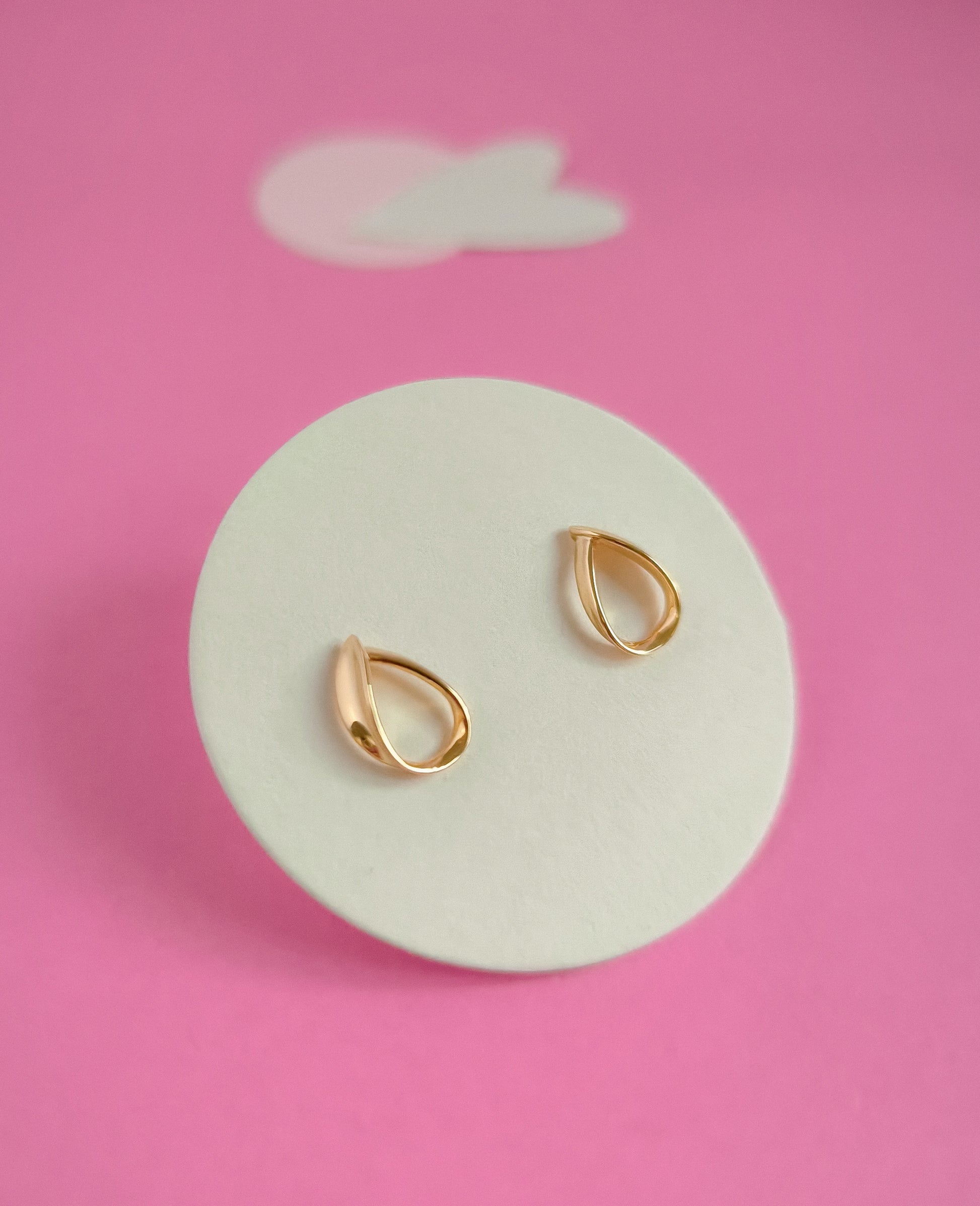 Gold teardrop earrings on a white circular stand against a pink background