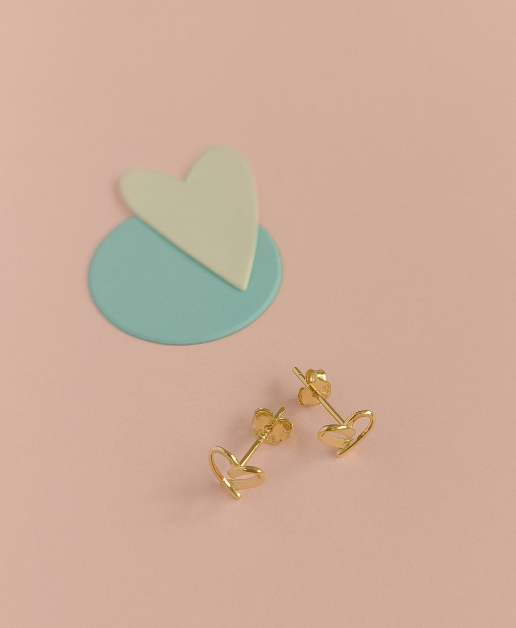 Gold heart-shaped earrings on a pink background with a heart-shaped stand.