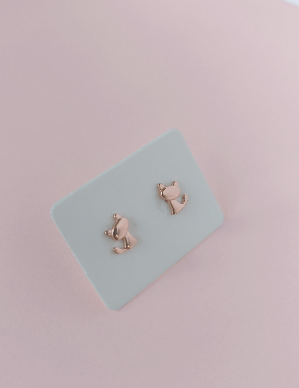 Rose gold cat-shaped earrings on a light blue card with a pink background