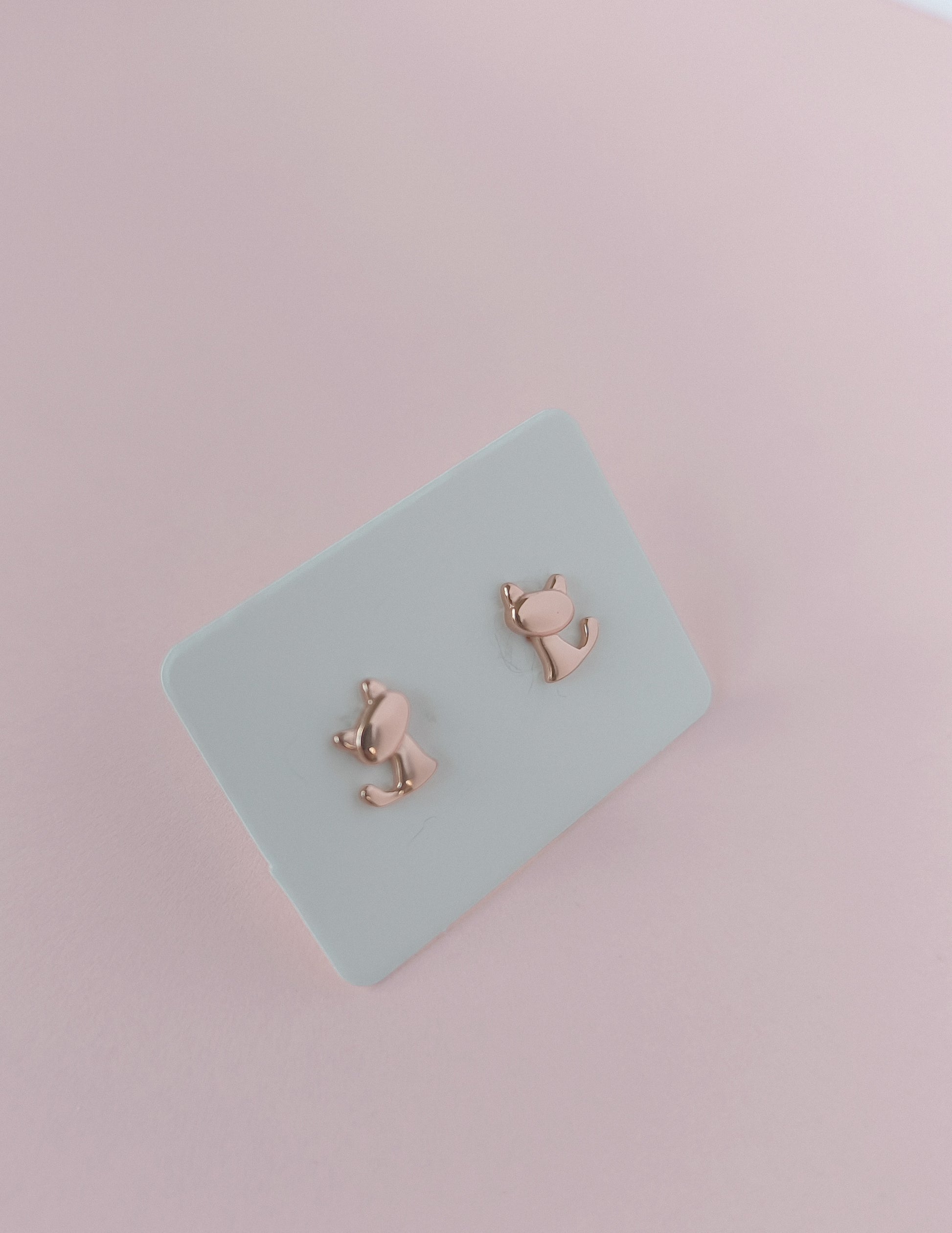 Rose gold cat-shaped earrings on a light blue card with a pink background